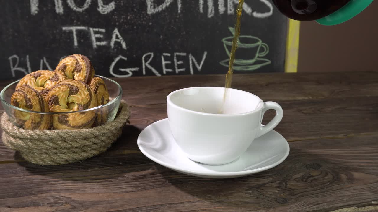 una taza de té y galletas caseras en una mesa de café. el té está derramando de una tetera transparente.