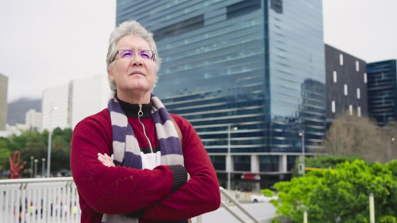 Ein älterer Mann lehnt in rotem Pullover an einem Geländer am Gehweg, bemerkt ein gläsernes Hochhaus und überblickt die Skyline.