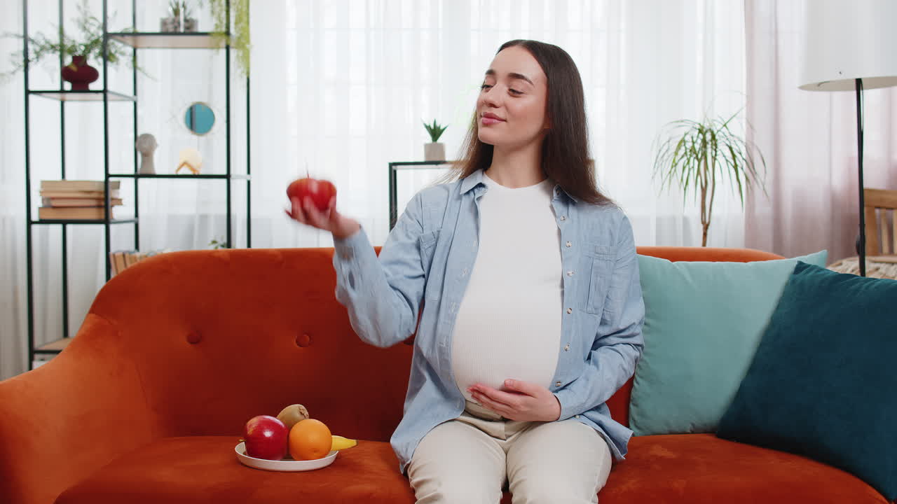 Happy pregnant woman eating fresh red apple fruit sitting on sofa healthy vitamin food motherhood
