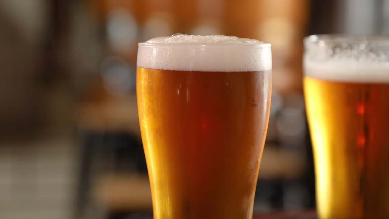 Un primo piano mostra due boccali di birra con schiuma in superficie. Un boccale è perfettamente a fuoco, mentre l'altro è sfocato sullo sfondo. L'ambiente suggerisce un bar o un pub.