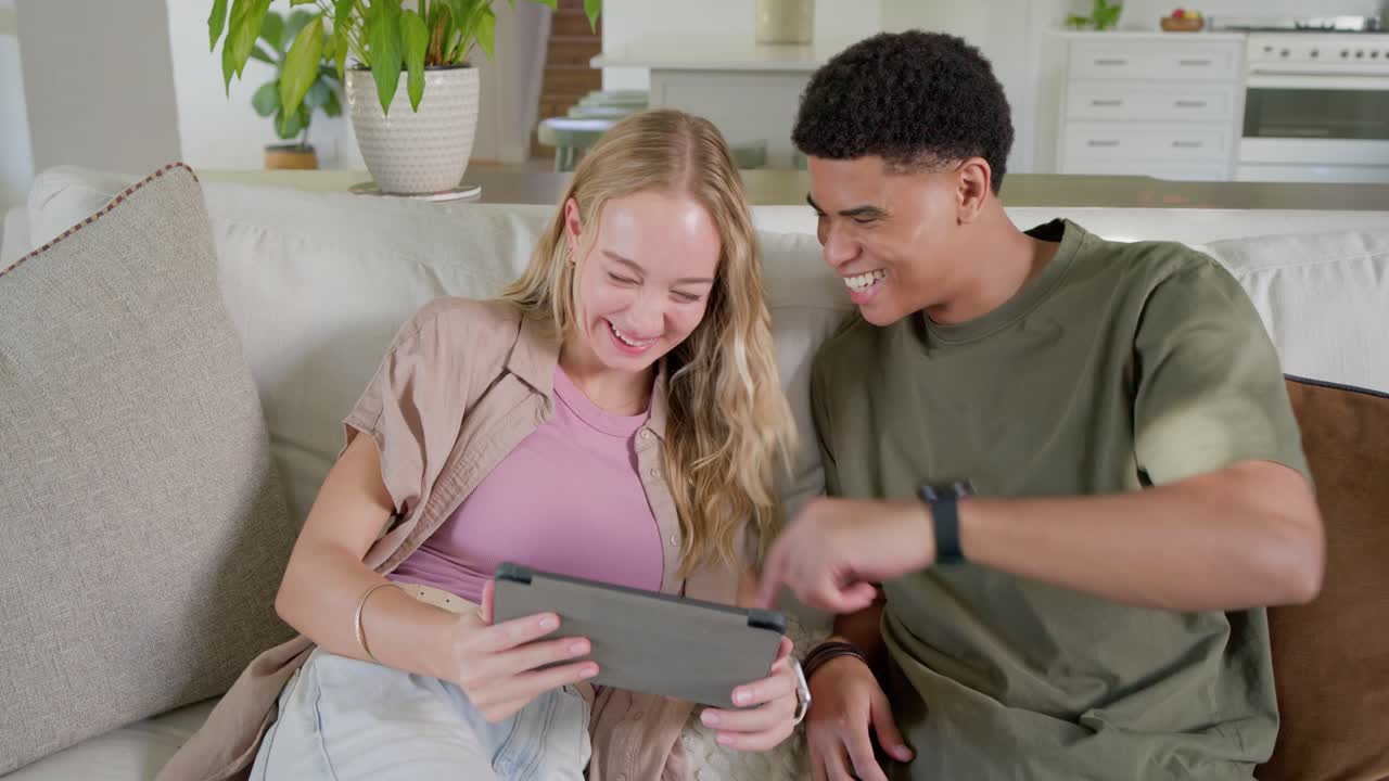 Couple sitting on light sofa in bright living room, watching funny clip on tablet, laughing