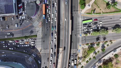 fotografía aérea descendente que muestra el tráfico urbano con vehículos que cruzan en varias direcciones, estructuras elevadas visibles en la distancia, todas iluminadas por la luz del día en una intersección concurrida