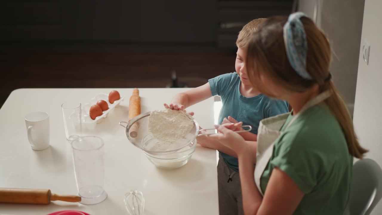 overhead view woman holds sieve while child taps flour into glass bowl on white counter surrounded by eggs rolling pin whisk cookie cutters capturing playful baking lesson moment in kitchen