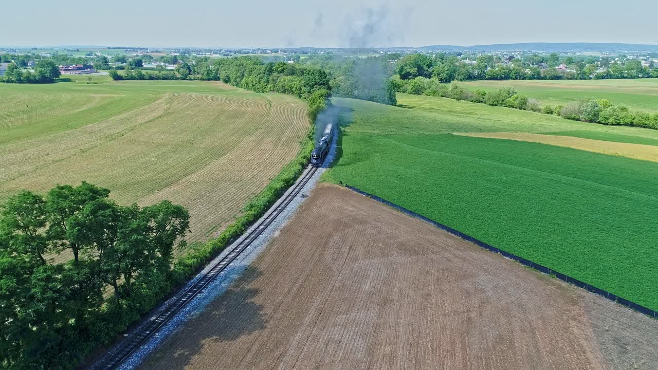 An Aerial View of a Steam Engine Puffing Smoke and Steam with Passenger Coaches Traveling on a Single Track Thru Trees and Farmland Countryside on a Beautiful Cloudless Spring Day