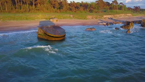 Aerial-view-of-abandoned-seaside-fortification-buildings-at-Karosta-Northern-Forts-on-the-beach-of-Baltic-sea-,-waves-splash,-golden-hour-sunset,-wide-drone-shot-moving-forward