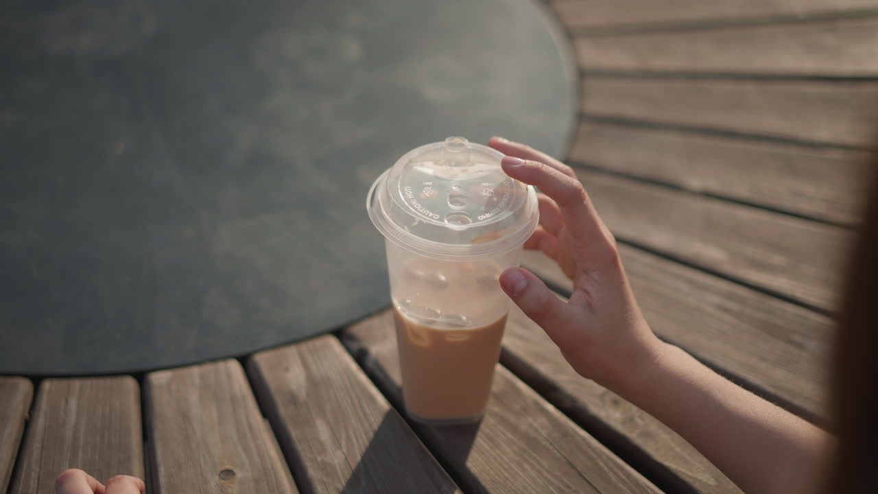 Tavolo dorato in legno con caffè freddo, mano che si allunga e spalla sfocata che entra nell'inquadratura, movimento catturato in un morbido effetto bokeh, contrasto di ombre calde, interazione spontanea su una terrazza urbana, invito a un sorso casual