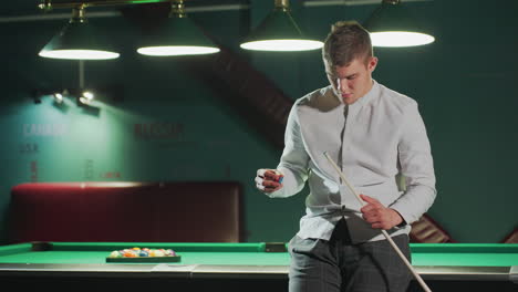 player in white shirt and grey trousers sits on green pool table, holding cue stick and carefully applying red cover with a focused expression. overhead lighting highlights