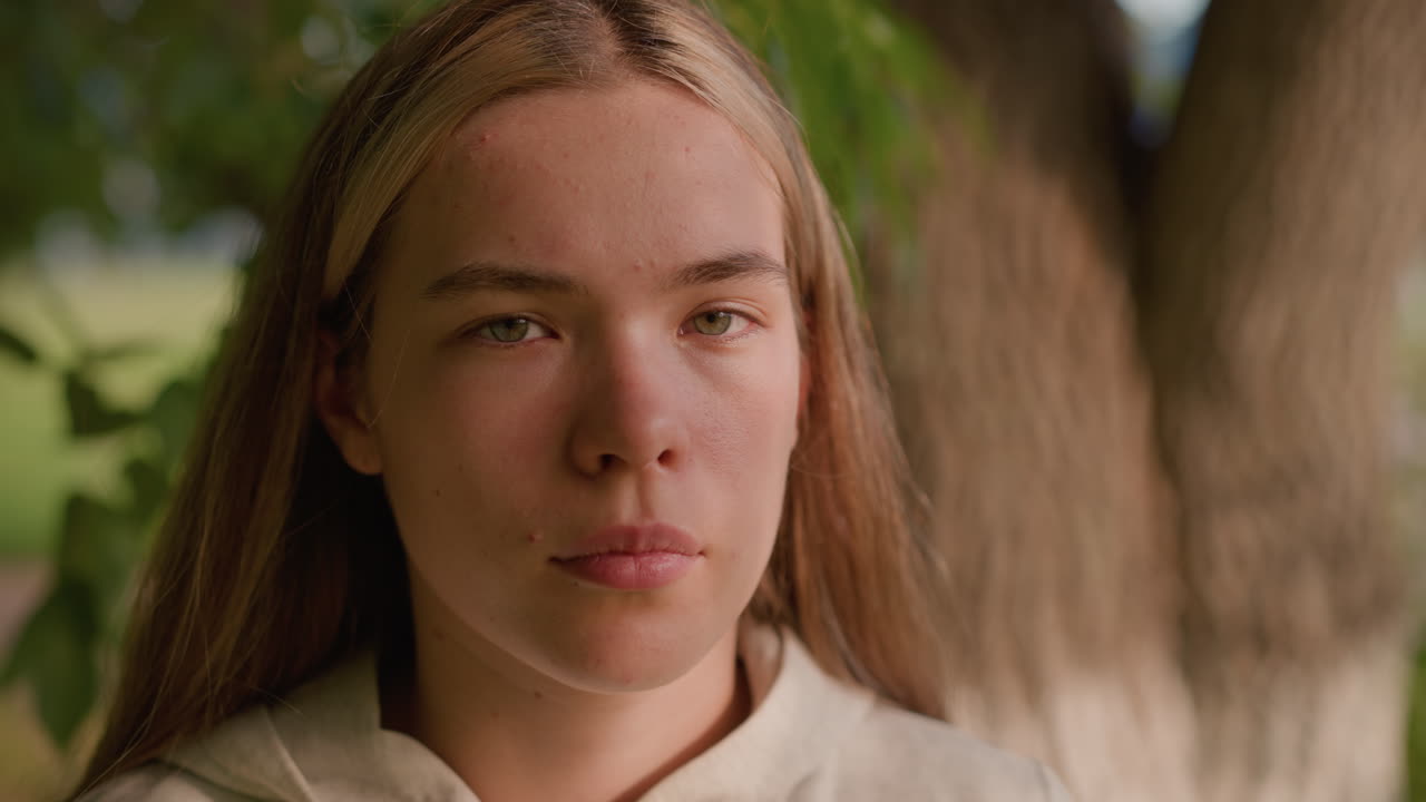 молодая леди с сосредоточенным выражением лица, стоящая на открытом воздухе с спокойной интенсивностью в естественной обстановке, солнечный свет бросает мягкие тени на ее лицо, смешиваясь с фоной зелени