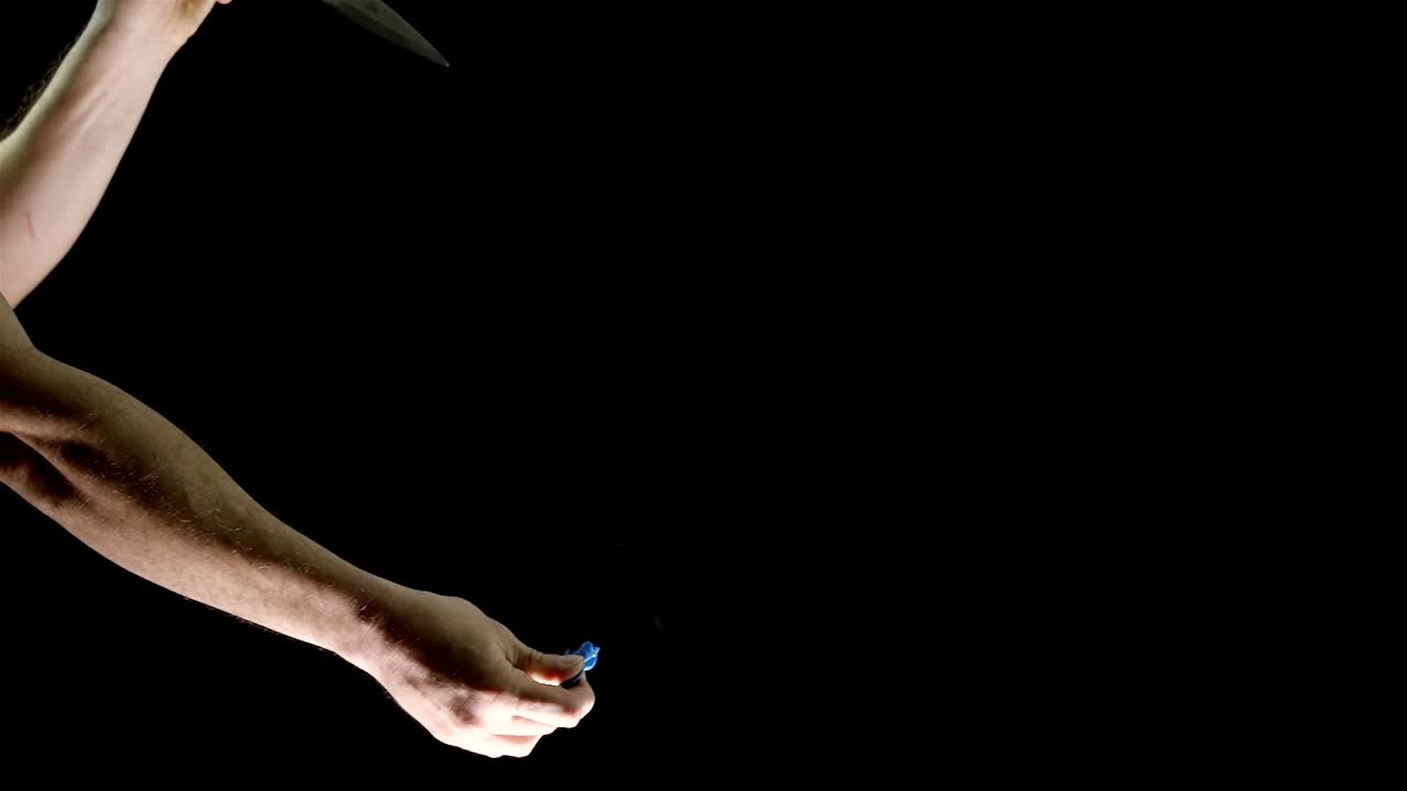 Watch as a blue balloon is held by one hand, while scissors in the other hand slice through the balloon, causing it to pop. This dramatic close-up captures the tension and excitement in slow motion.
