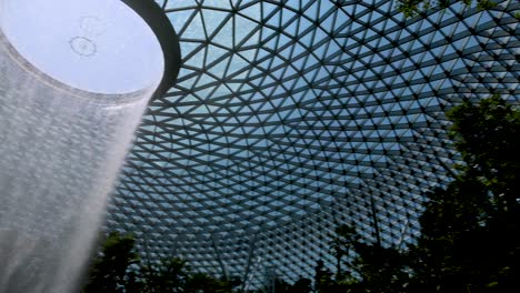 a dramatic indoor waterfall pours from a circular skylight in a vast glass-domed atrium, surrounded by vibrant greenery and natural daylight. slow upward camera movement
