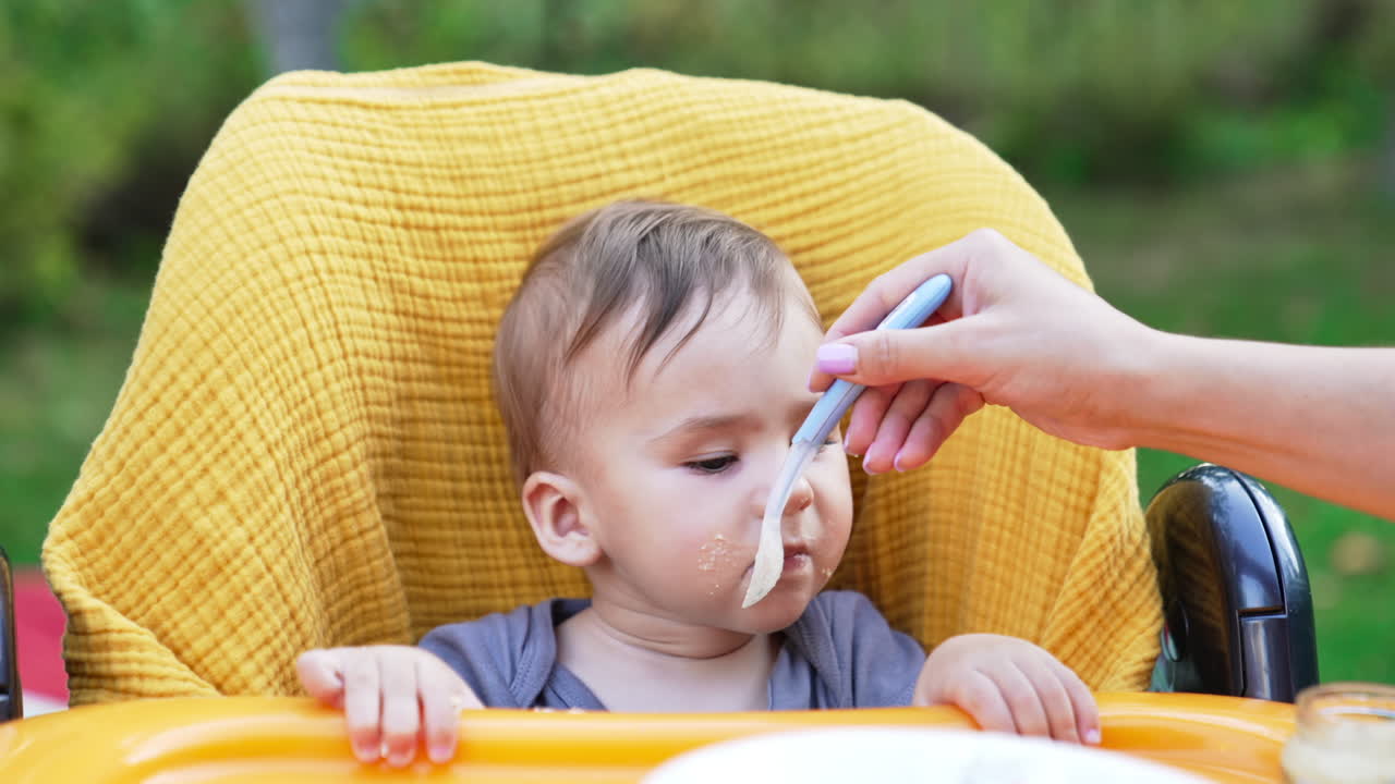 Sweet little baby boy sitting in the yellow chair. Cute kid with smudgy face eating porridge from spoon. Close up.
