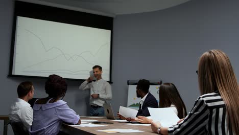 un joven empleado de la empresa con una camisa blanca informa sobre el progreso del trabajo realizado en el último año y los planes de desarrollo para el trimestre ocupado en el diagrama del proyector en la oficina. informe a la junta directiva un equipo multiétnico