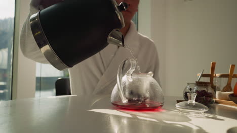 un hombre vierte agua en una tetera llena de hojas de té. un hombre vestido con túnica de baño prepara una bebida caliente sentado en la mesa de comedor. preparando una bebida contra la vista de la naturaleza
