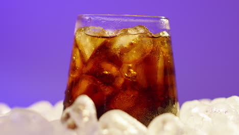 Close-Up-Of-Chilled-Cold-Drink-In-Glass-Being-Mixed-On-Ice-Cubes-Against-Purple-Background