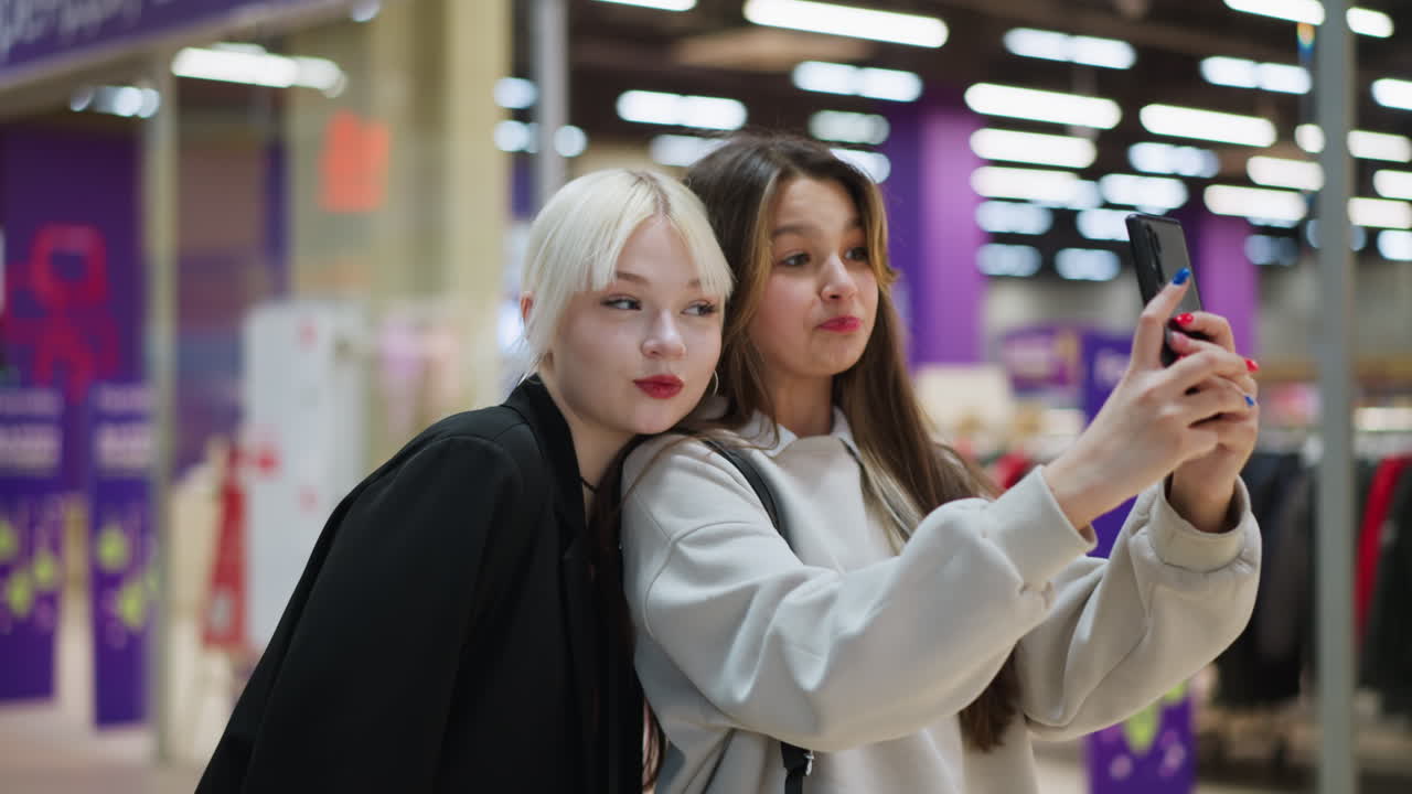 Two students use smartphone while walking inside modern shopping mall, smiling and looking at screen together under bright ceiling lights with colorful retail environment in background