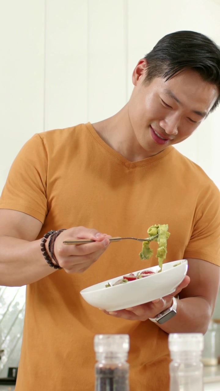 垂直影片：一名穿橙色T恤的男子在廚房裡拿起白色碗，用叉子攪拌並檢查沙律。