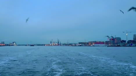 wide shot of amsterdam's industrial ij harbor with construction cranes and modern waterfront buildings. the soft blue evening light and water create a tranquil yet bustling urban landscape