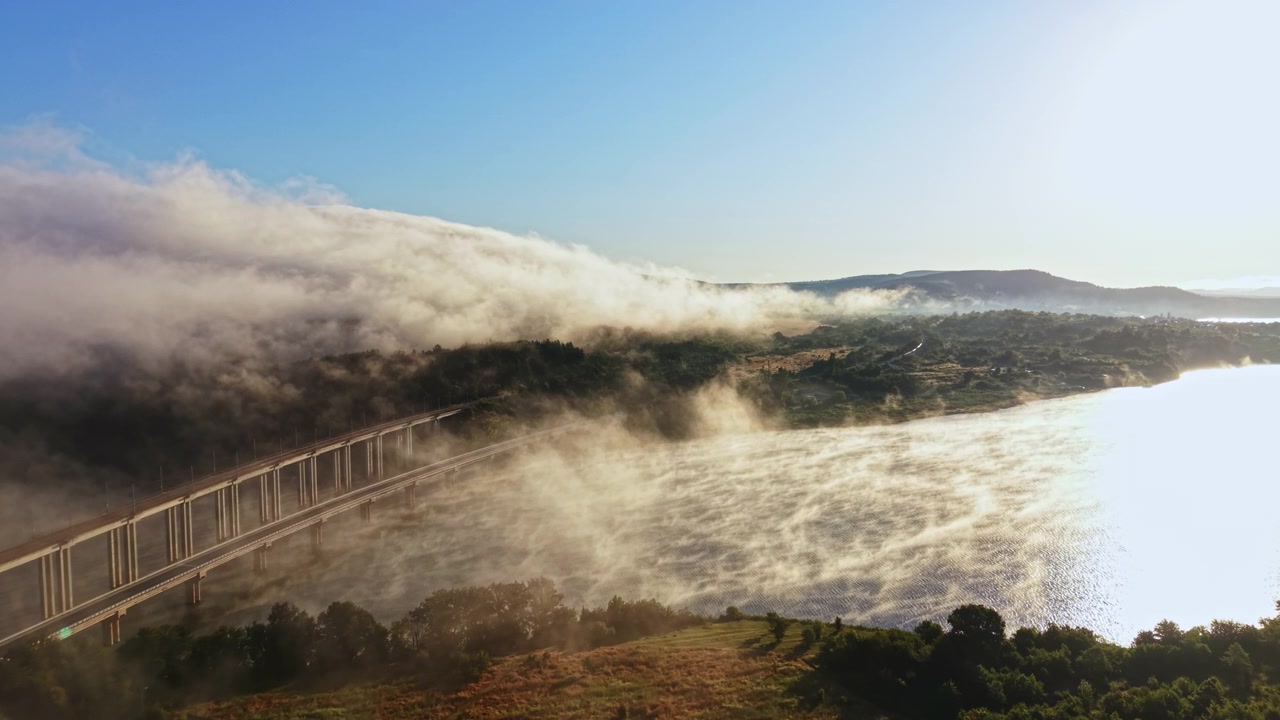 Vista de neblina sobre um rio perto de uma ponte durante as primeiras horas da manhã com a luz do sol rompendo as nuvens.