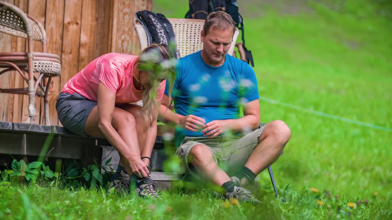 An older man and a blonde woman who is tying her shoelace, getting ready for their hiking trip in a green valley