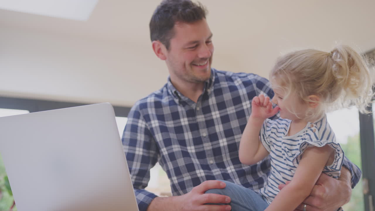 padre trabajando usando una computadora portátil en casa en el mostrador de la cocina mientras cuida a su hija pequeña - filmado en cámara lenta