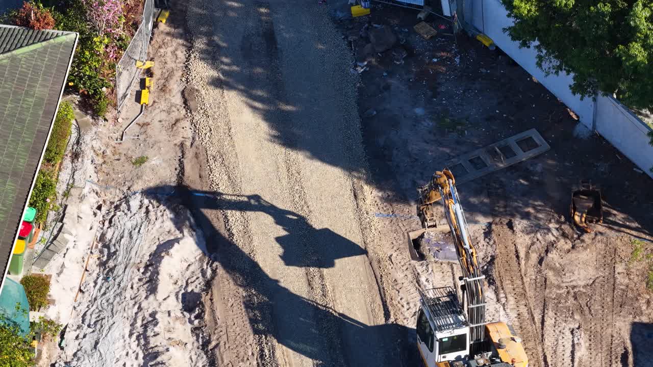 Aerial view of excavator leveling dirt road during sunny daytime construction in residential neighborhood