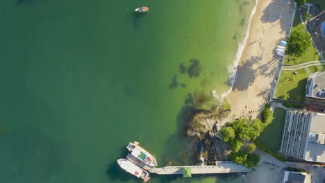 Aerial-top-view-panning-over-the-shore-of-a-brazilian-paradise-beach-with-white-sand,-emerald-clear-water,-boats-and-rocks