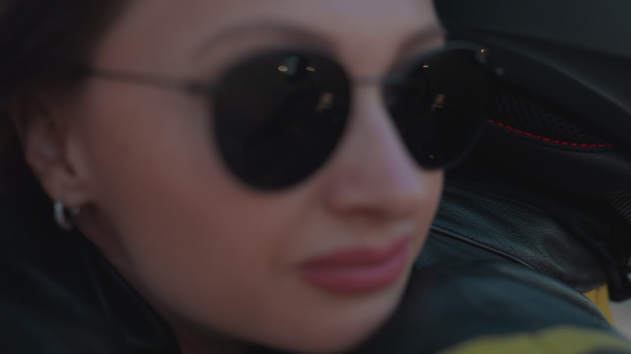 Close up of woman in leather stepping onto motorcycle while holding rider, sunlight creating warm glow with billboard and urban buildings blurred in background