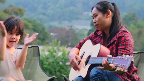 mother and child play guitar and sing together on camping chairs near tent at camp in summer forest. the family spends time together on vacation.