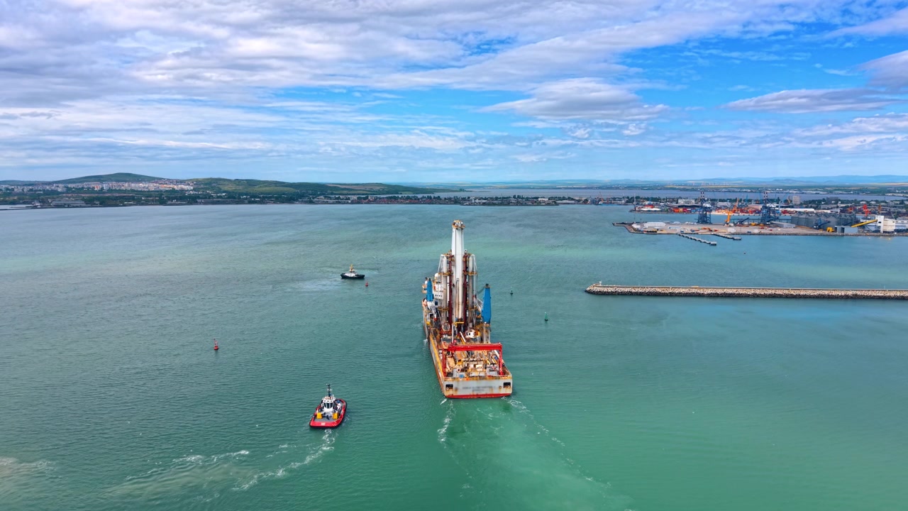 Luftfoto af offshore boreskib eskorteret af slæbebåd. Droneoptagelse af industriboreskib med boretårn og slæbebåd på turkisblåt hav med kommerciel havn i baggrunden.