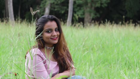 beautiful indian woman sitting in tall grass field smiling then look up to the camera