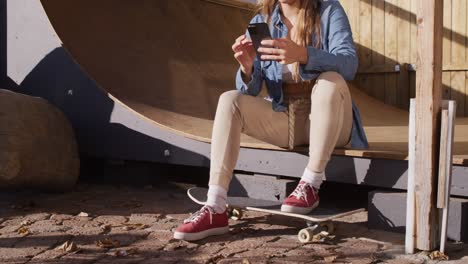 video of happy caucasian female skateboarder resting and using smartphone