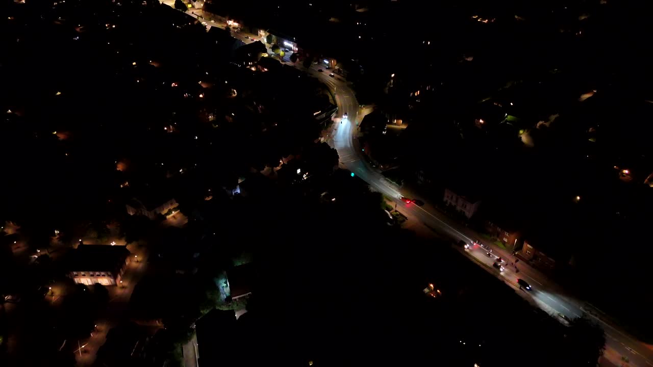 Driving cars on motorbikes on illuminated street of small American town. Aerial Birds Eye shot. Night scene in America neighborhood with curvy Main Street. Roundabout in background