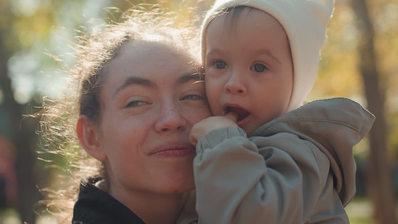 Nahaufnahme einer Mutter, die ihr Baby in einem sonnendurchfluteten Park küsst. Herbstliche Wärme und zärtliche Familienzuneigung. Junge Mutter hält Kleinkind eng bei sich, weiches goldenes Licht. Gemütliche Mütze und Jacke. Ungezwungenes Porträt, das mütterliche Zuneigung einfängt.