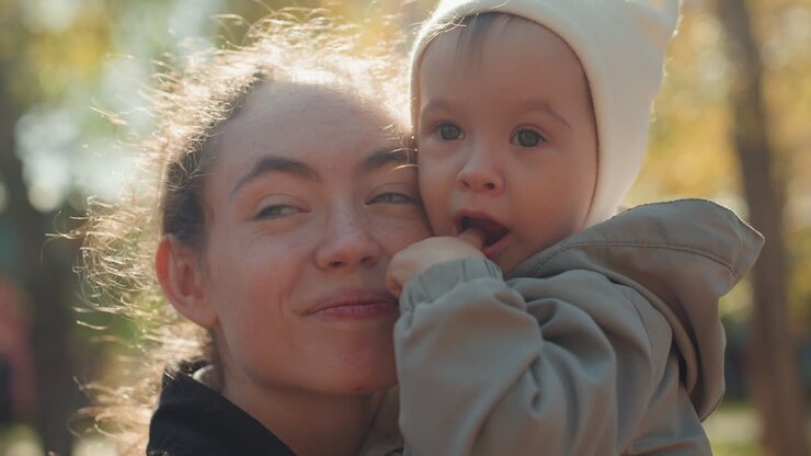 Nahaufnahme einer Mutter, die ihr Baby in einem sonnendurchfluteten Park küsst. Herbstliche Wärme und zärtliche Familienzuneigung. Junge Mutter hält Kleinkind eng bei sich, weiches goldenes Licht. Gemütliche Mütze und Jacke. Ungezwungenes Porträt, das mütterliche Zuneigung einfängt.