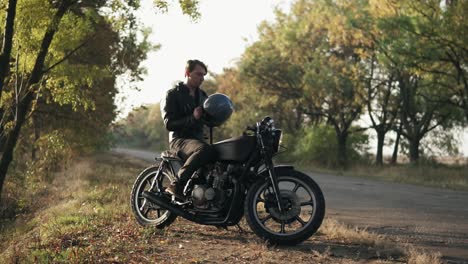 a young man in black leather jacket sits on his motorcycle and puts a black helmet to go on a journey in the forest. slow motion