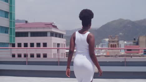 a black girl in a white dress walking on a rooftop with the city skyline in view