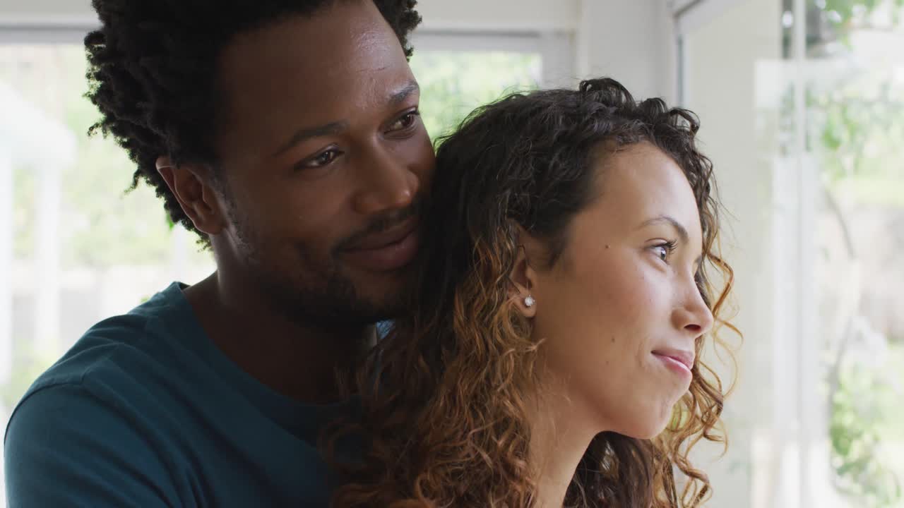 Profiles of happy biracial couple embracing, standing at window and looking into distance