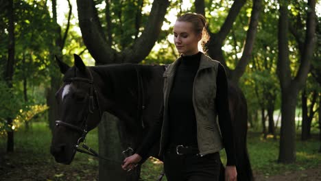 disparo en cámara lenta de una joven y atractiva jockey caminando con un caballo marrón con una mancha blanca en la frente en el bosque durante el sol