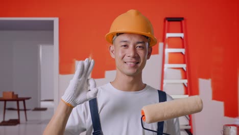 primer plano de un pintor asiático que lleva un casco de seguridad sonriendo y mostrando un gesto de acuerdo a la cámara mientras está de pie con la pared de pintura en la habitación