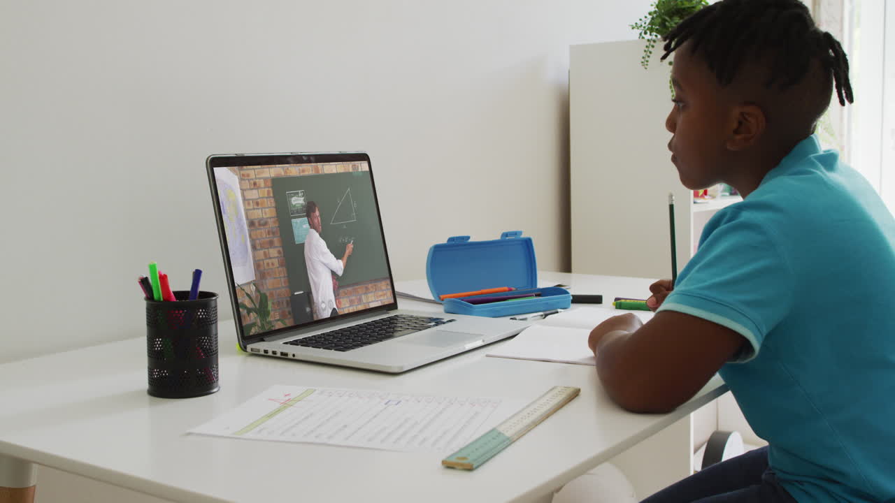 niño afroamericano haciendo la tarea mientras tiene una videollamada con un profesor masculino en una computadora portátil en casa