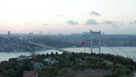 Türkische-Flagge-Weht-Im-Wind-Vor-Der-Bosporus-Brücke-In-Istanbul-Und-Der-Skyline-Der-Stadt-Bei-Schönem-Sonnenuntergang-Und-Richtet-Die-Luftrutsche-Nach-Rechts-Ein