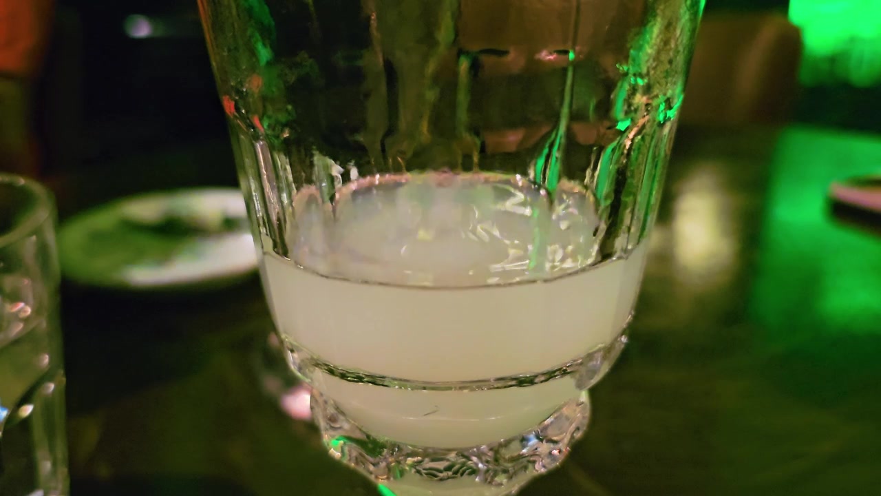 Primer plano extremo de absenta turbia dentro de una copa de cóctel, con líquido blanco cremoso, detalles de vidrio texturizado, reflejos de mesa oscura e iluminación verde de vida nocturna en un bar tenue.