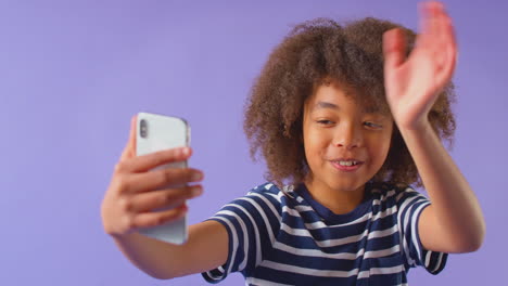 retrato de estudio de un niño agitando haciendo una llamada de video en un teléfono móvil contra un fondo púrpura