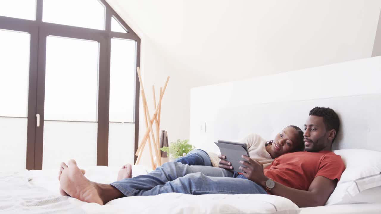 Happy african american couple lying on bed using tablet, in slow motion