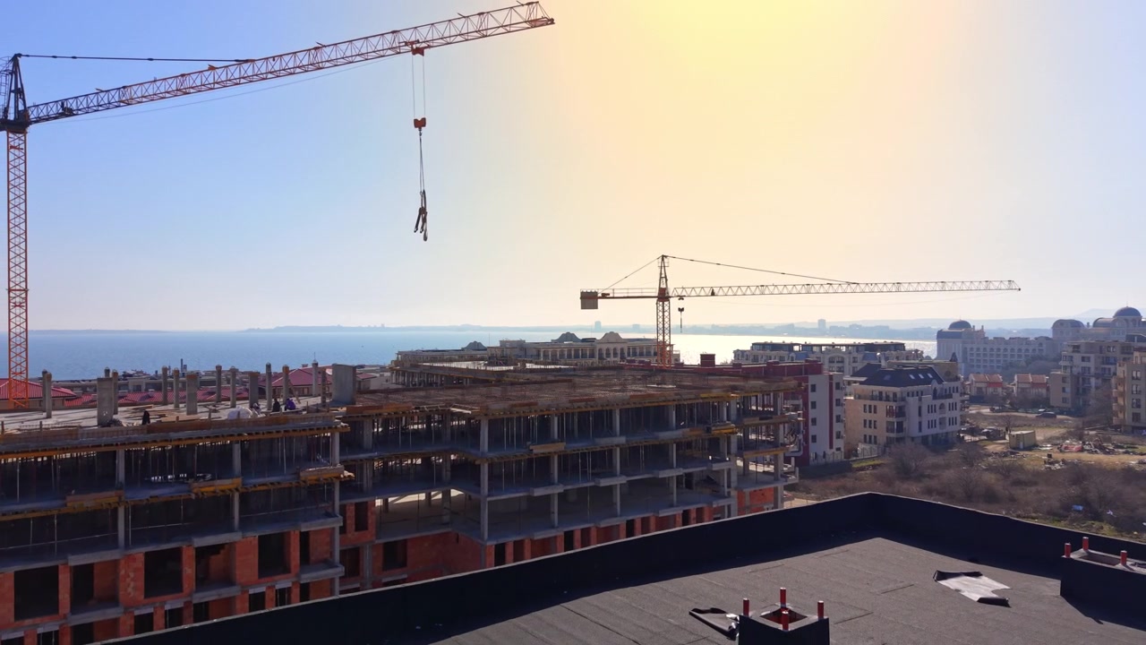 Aerial view of crane hook over coastal construction site, Bulgaria. Hazy sunlight glows above unfinished apartment building and rooftops near the sea.