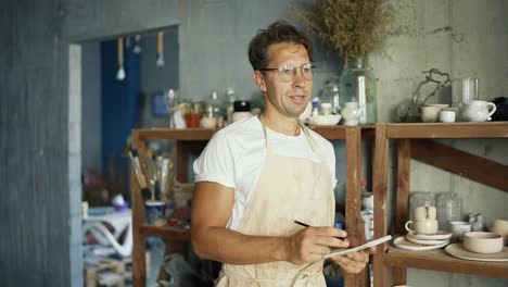 the young potter is doing inventory looking at ceramics and taking notes in workshop. the potter writes down his plans for future