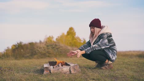 una mujer excursionista calienta las manos en una hoguera en el campamento en otoño