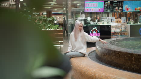 blurred leafy foreground shows shopper seated by illuminated water fountain inside busy mall, manicured hand skimming clear flowing water while neon bubble tea kiosk glows in vibrant background