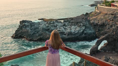 high angle view of woman looking out to coastal sea cliffs, waves crash dreamily in slow motion