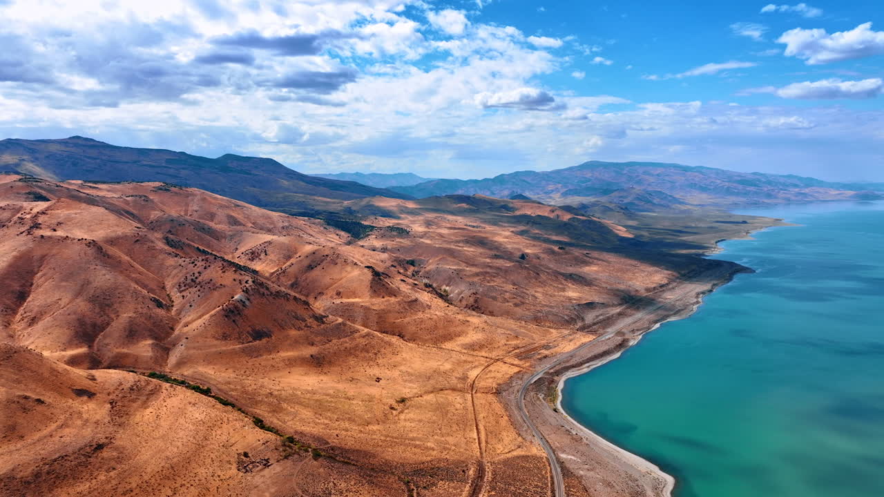 Bare mountainous landscape and a waterscape at the foot. Highway passes along the waterfront. Fluffy clouds in the azure sky